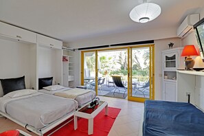 Hotellihuone, jossa on kaksi sänkyä, punainen matto, tarjotinalustalla varustettu pöytä, tuoli, lamppu ja näkymä ulos liukuovien kautta.