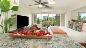 Una sala de estar moderna con un televisor de pantalla plana, una mesa de centro de madera con fruta, un ventilador de techo y un balcón con vista a la ciudad.