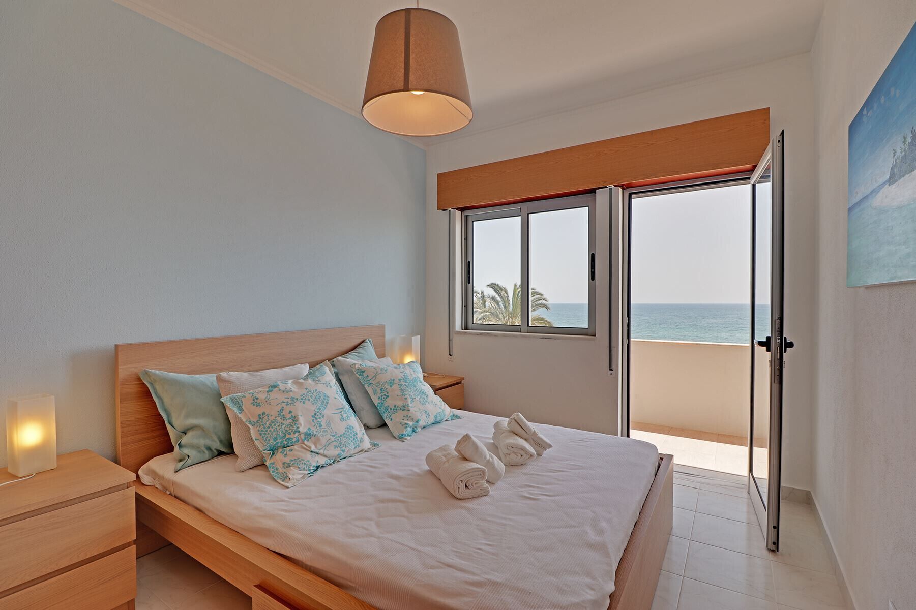 Un dormitorio con una cama de madera, una mesita de noche, una lámpara de noche, una ventana con vistas al mar y un cuadro en la pared.