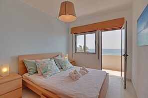 A bedroom with a wooden bed, a nightstand, a bedside lamp, a window with a view of the sea, and a painting on the wall. - Quarteira Beach & Ocean View 1 by Homing (Loulé)