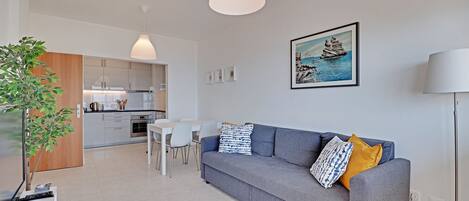 A modern living room with a grey sofa, a dining table, and a kitchen in the background.
