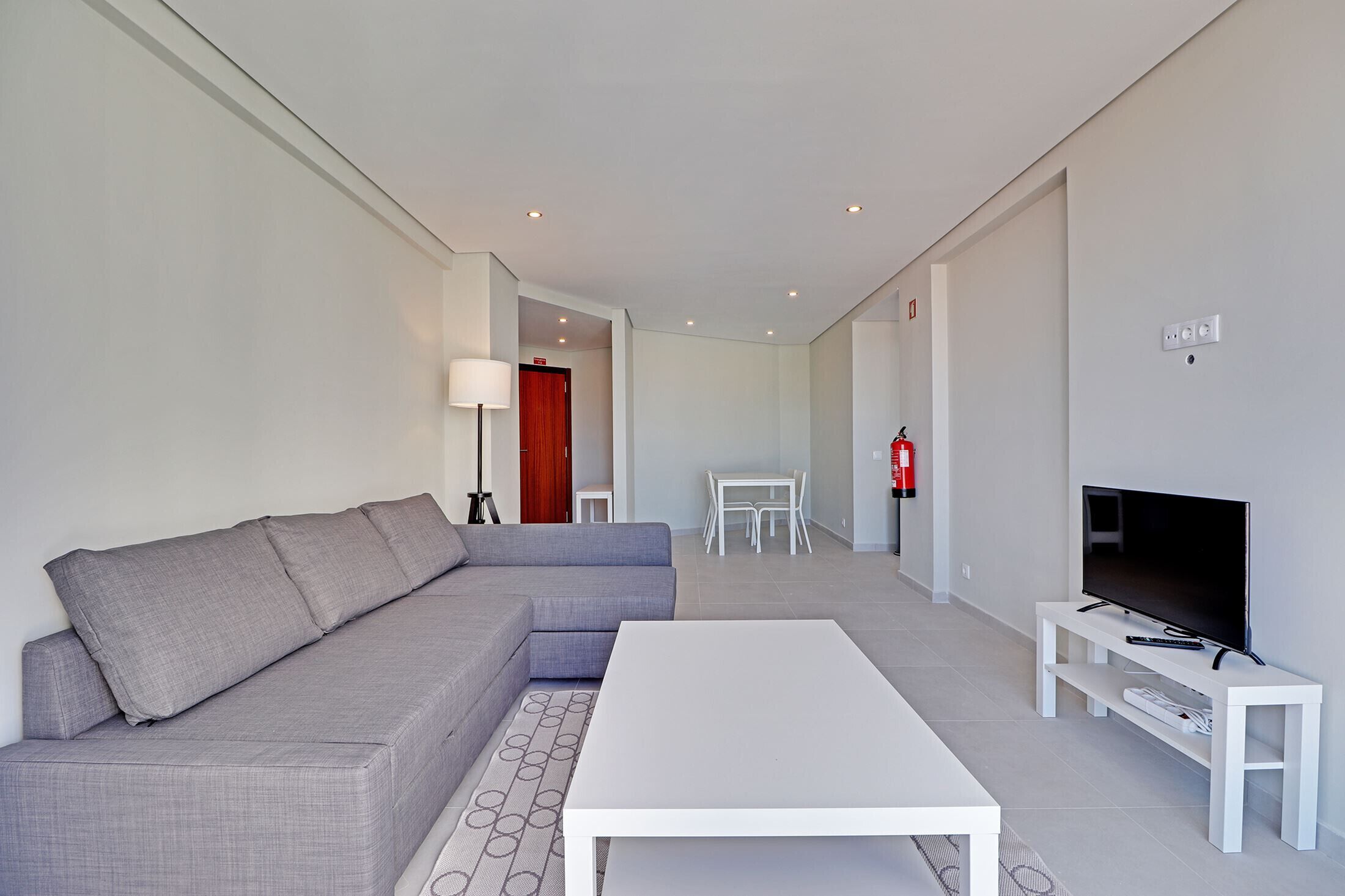 Un salon moderne avec un canapé d’angle gris, une table basse blanche et un téléviseur posé sur un meuble blanc.