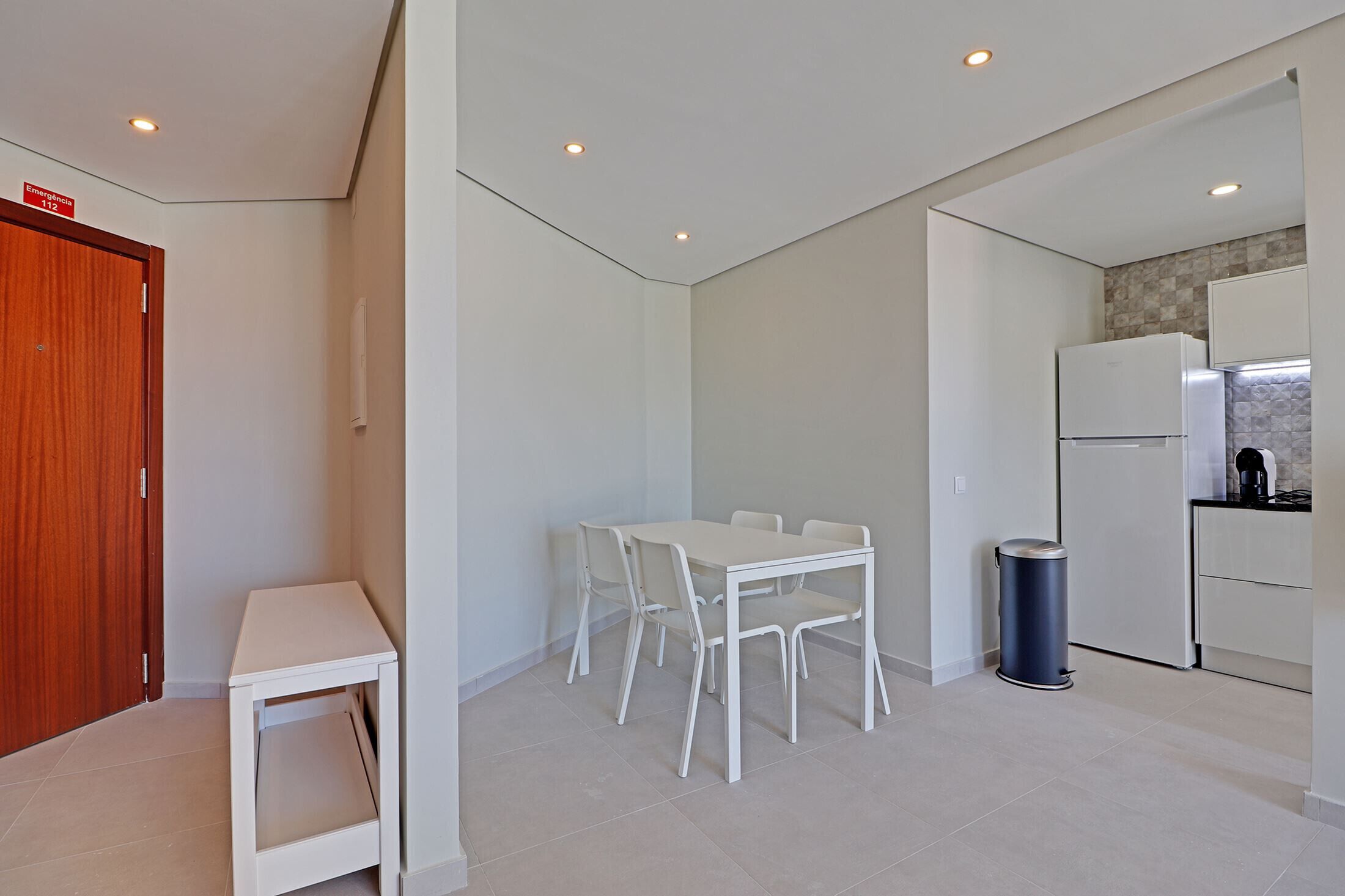 A modern kitchen with white cabinets, a built-in oven, and a refrigerator. There is a small dining table with chairs and a trash can.