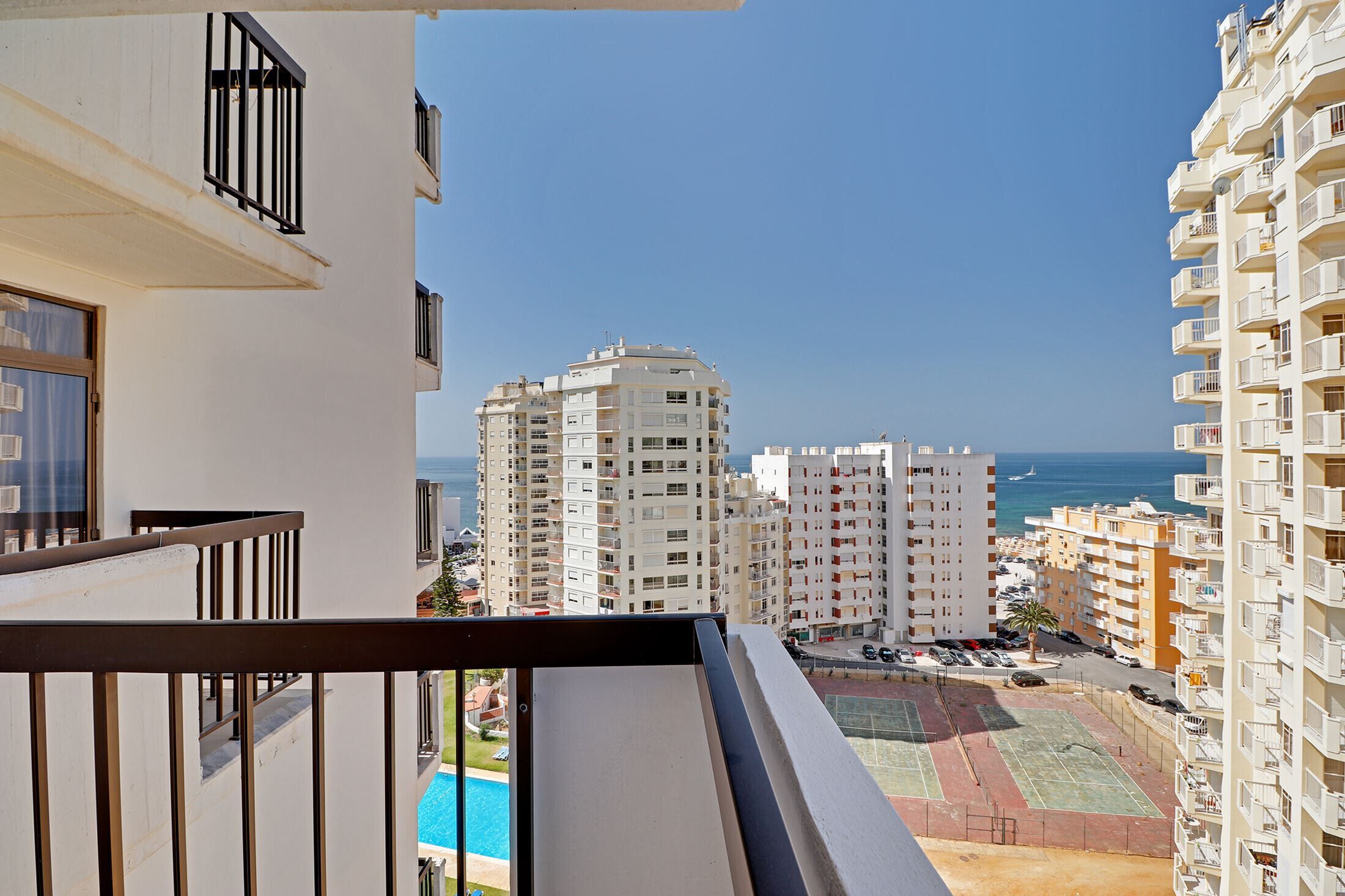 Un edificio alto con balconi che si affaccia su una piscina e campi da tennis, con vista sul mare e altri edifici sullo sfondo.