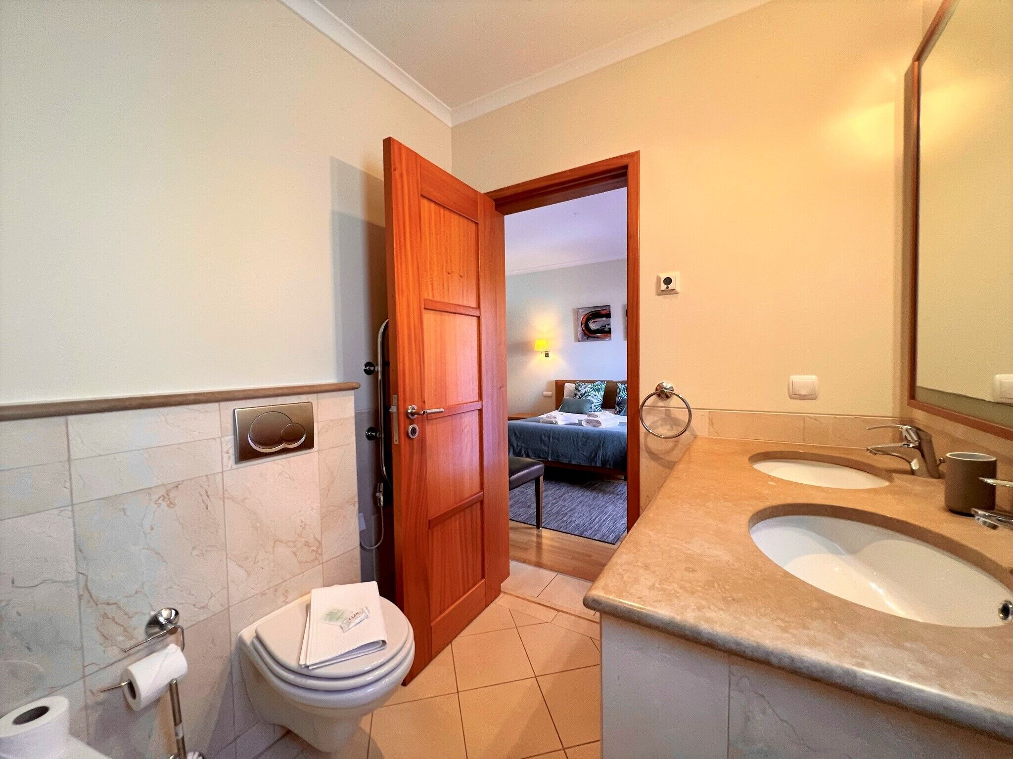 A bathroom with two sinks, a toilet, and a wooden door leading to a bedroom.