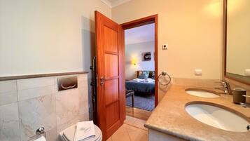 A bathroom with two sinks, a toilet, and a wooden door leading to a bedroom.