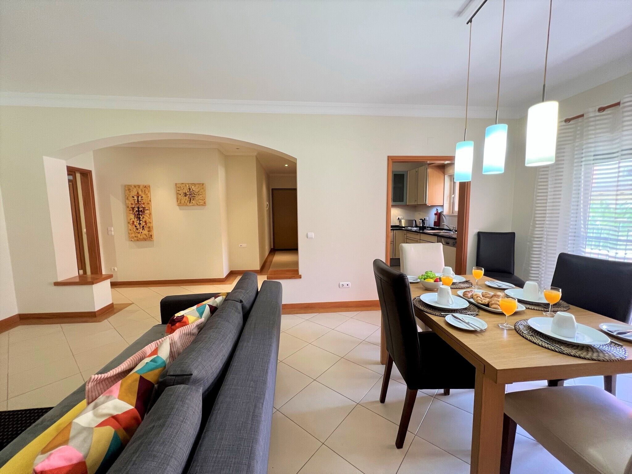 A modern dining area with a wooden table set for breakfast, a sofa with colorful cushions, and a kitchen in the background.