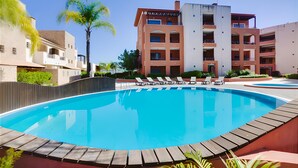 Una piscina amb gandules, envoltada per una zona verda amb palmeres i un edifici de diversos pisos al fons.