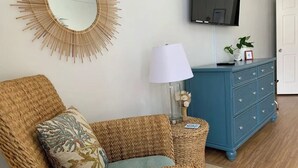 A room with a wicker chair, a blue dresser, a wall-mounted TV, and a mirror.