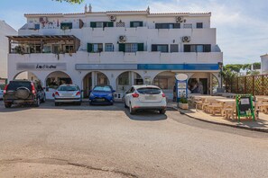 Un edificio con varias fachadas comerciales, un estacionamiento con varios coches y mesas al aire libre.