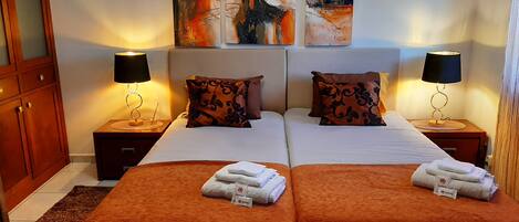 A double room with two beds, each with a brown bedspread, white linens, and matching pillows. There are two bedside tables with lamps, and the walls are adorned with abstract art.