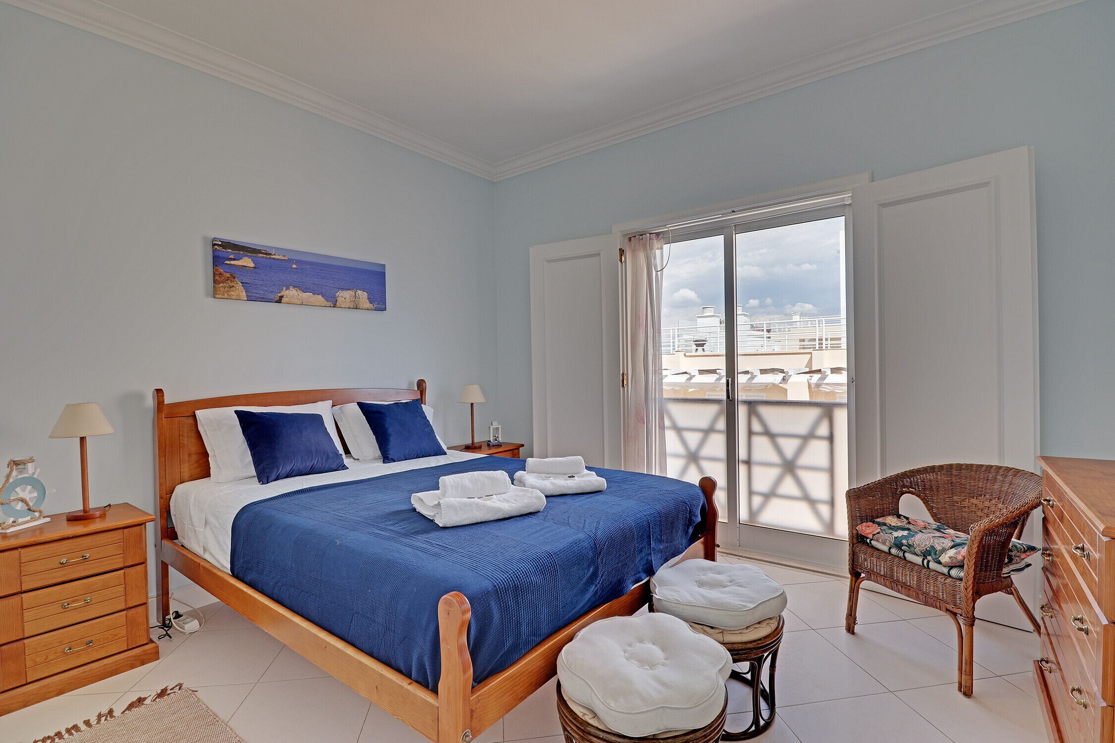 A bedroom with a wooden bed, a wicker chair, and a view of the sea through a balcony door.