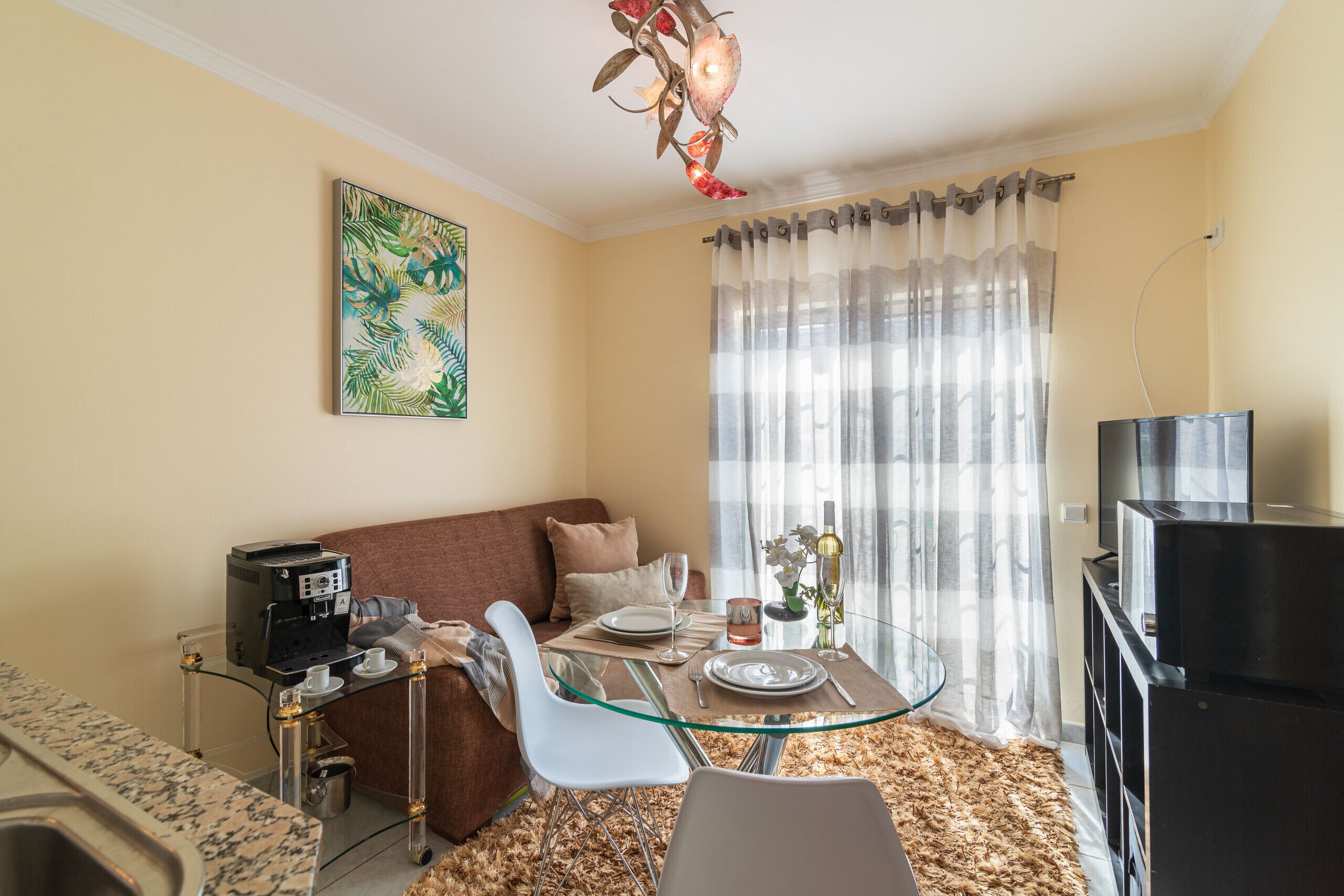 A compact living space with a glass dining table, a sofa, a TV, and a coffee machine.