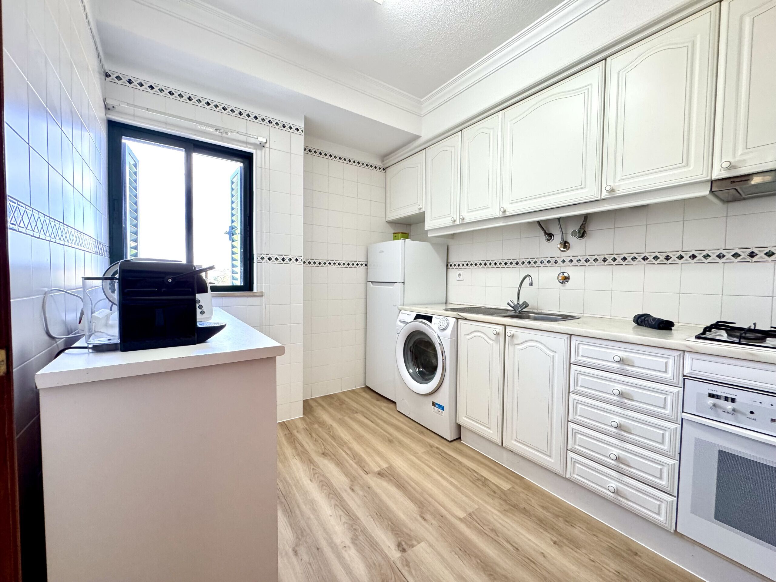 A compact kitchen with white cabinets, a built-in washing machine, a stove, and a window with blinds.