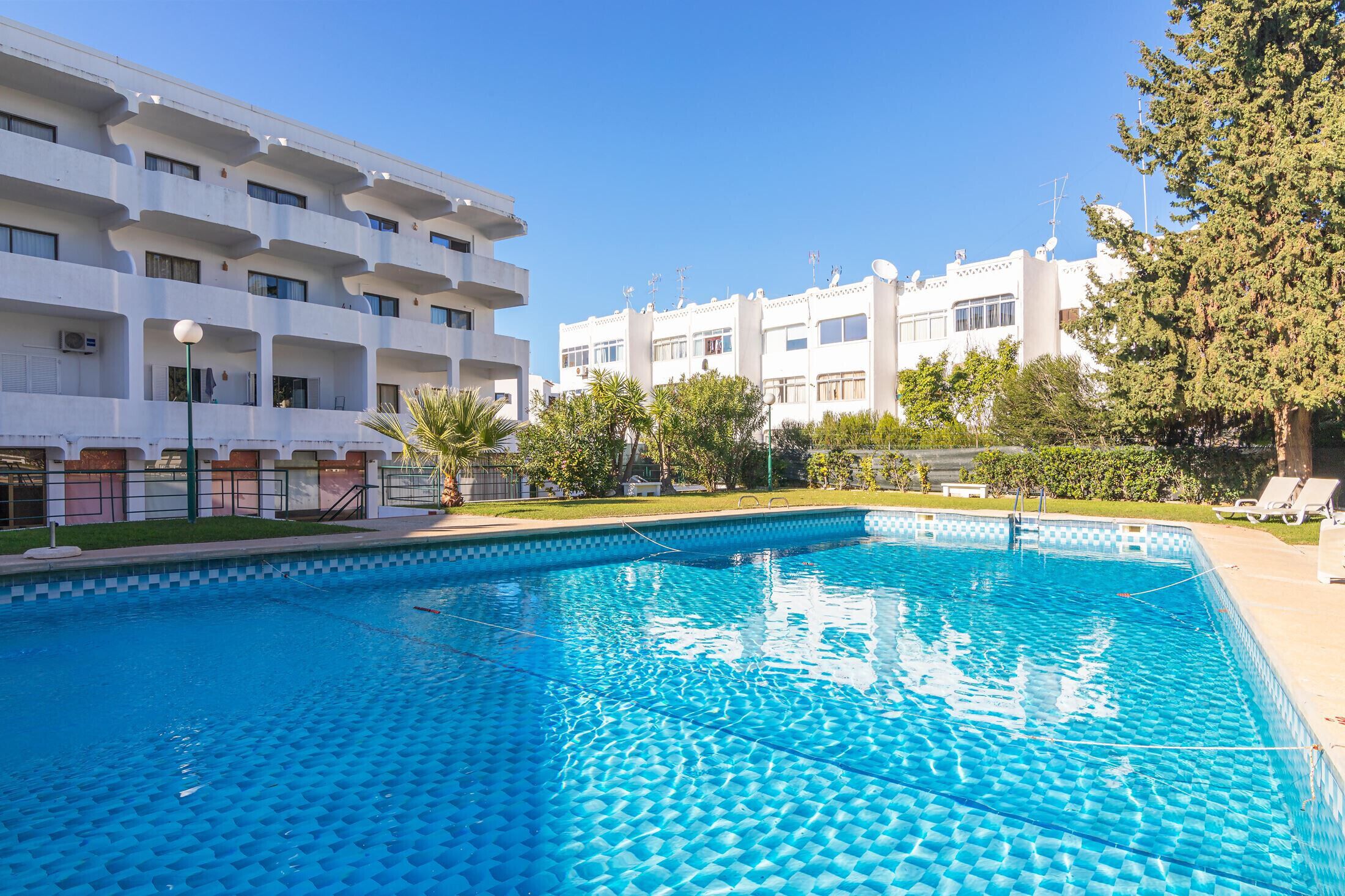 A swimming pool with a tiled deck, surrounded by a white building with multiple balconies and a clear blue sky.