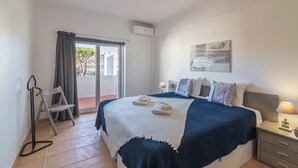 A hotel room with a bed, a nightstand, a chair, a balcony with a view, and a painting on the wall. - Vilamoura Cosy 2 With Pool by Homing (Loulé)