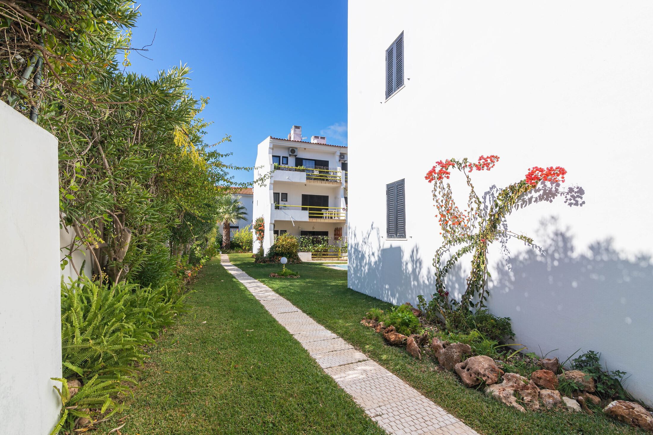 A pathway leading to a white building with a balcony and a garden with rocks and plants.