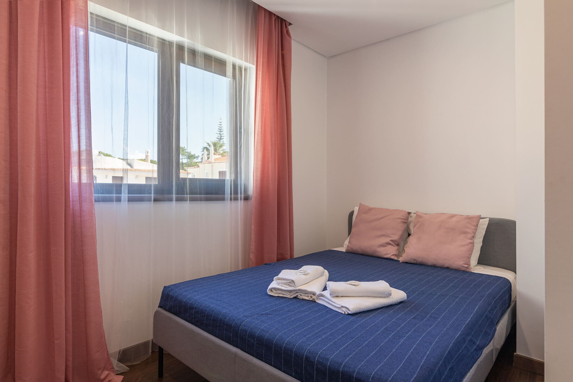 A hotel room with a bed, a window with sheer curtains, and a view of buildings outside.