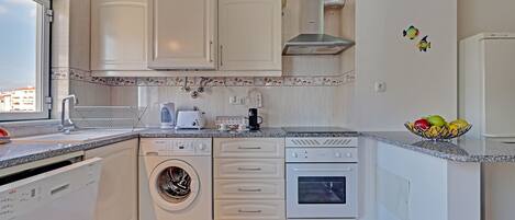 A compact kitchen with a dishwasher, washing machine, oven, and a small countertop with a fruit bowl.