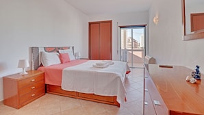 A bedroom with a bed, bedside table, and a dresser. There is a door leading to a balcony with a view of buildings. - Praia DA Rocha Central With Pool (Portimão)