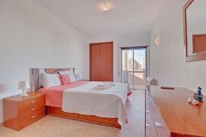 Ein Schlafzimmer mit Bett, Nachttisch und Kommode. Eine Tür führt zu einem Balkon mit Blick auf Gebäude.