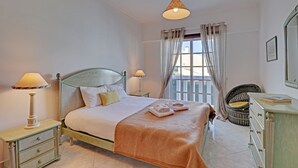 A neatly arranged bedroom with a bed, bedside lamps, a dresser, and a wicker chair. - Portimão Solar DO VAU With Pool (Portimão)
