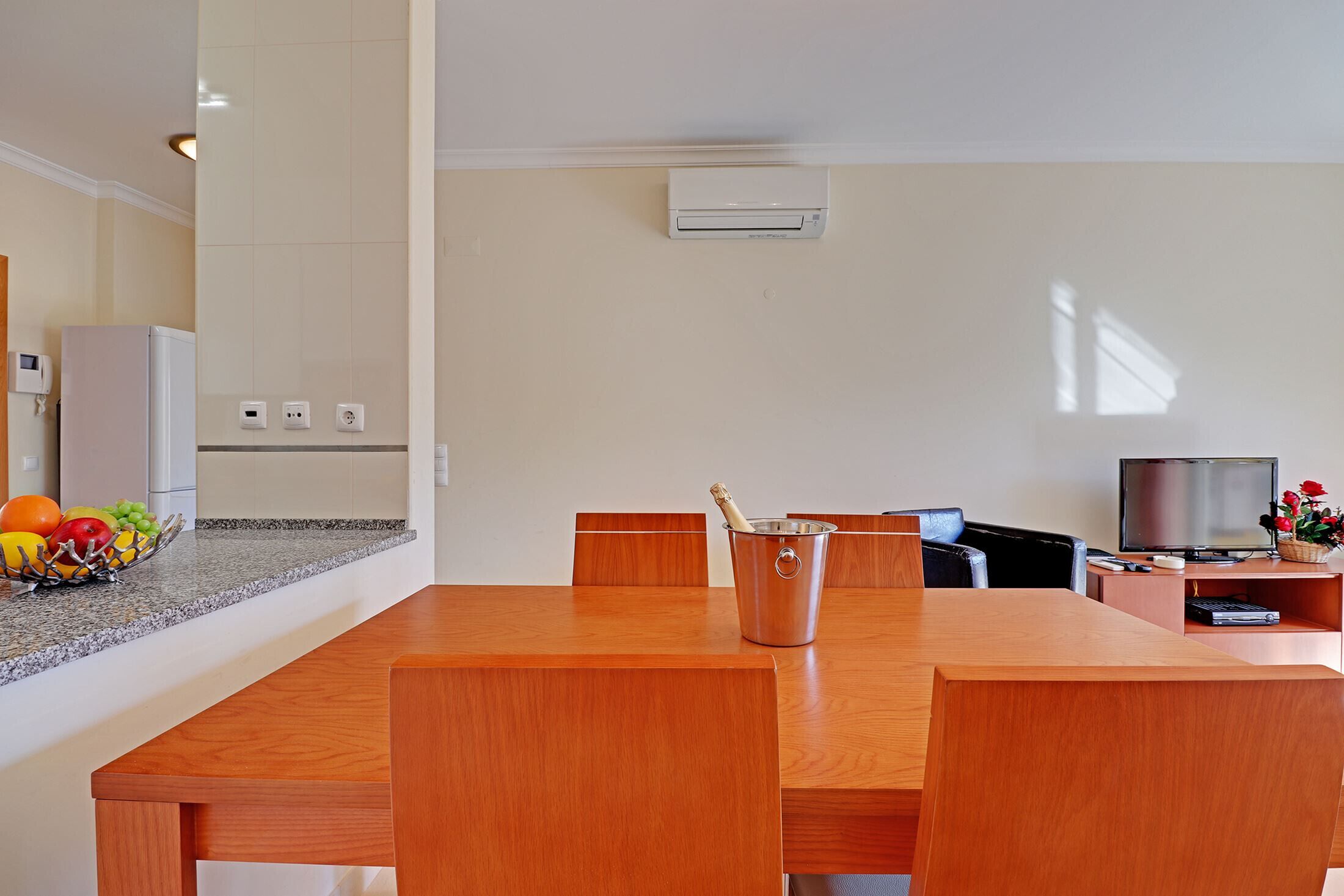 A modern kitchen with a built-in air conditioner, a dining area with a wooden table and chairs, and a television on a stand.