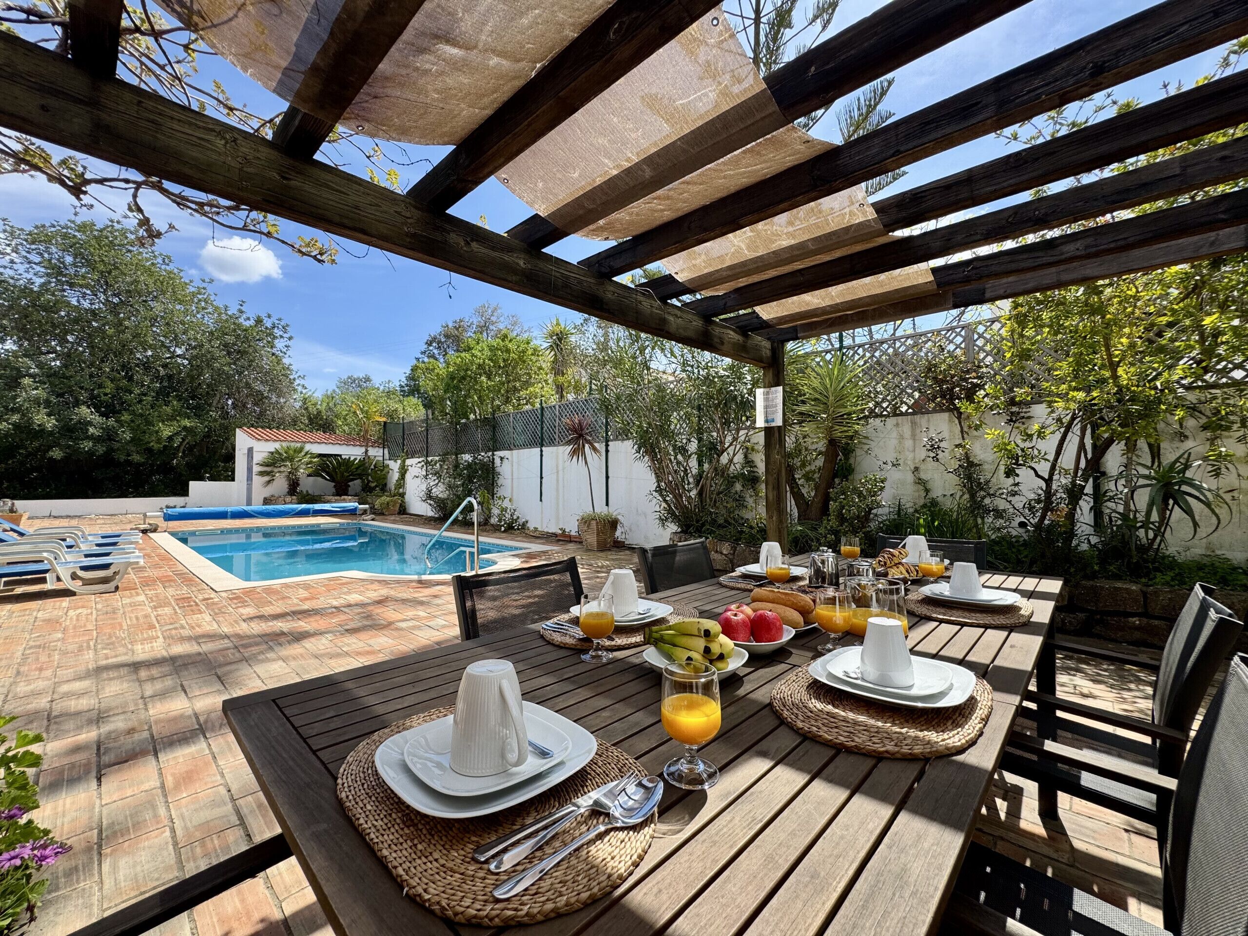 Eine Holzterrasse mit Pool, Essbereich im Freien mit Fruchtplatten und Getränken, sowie eine Pergola darüber.