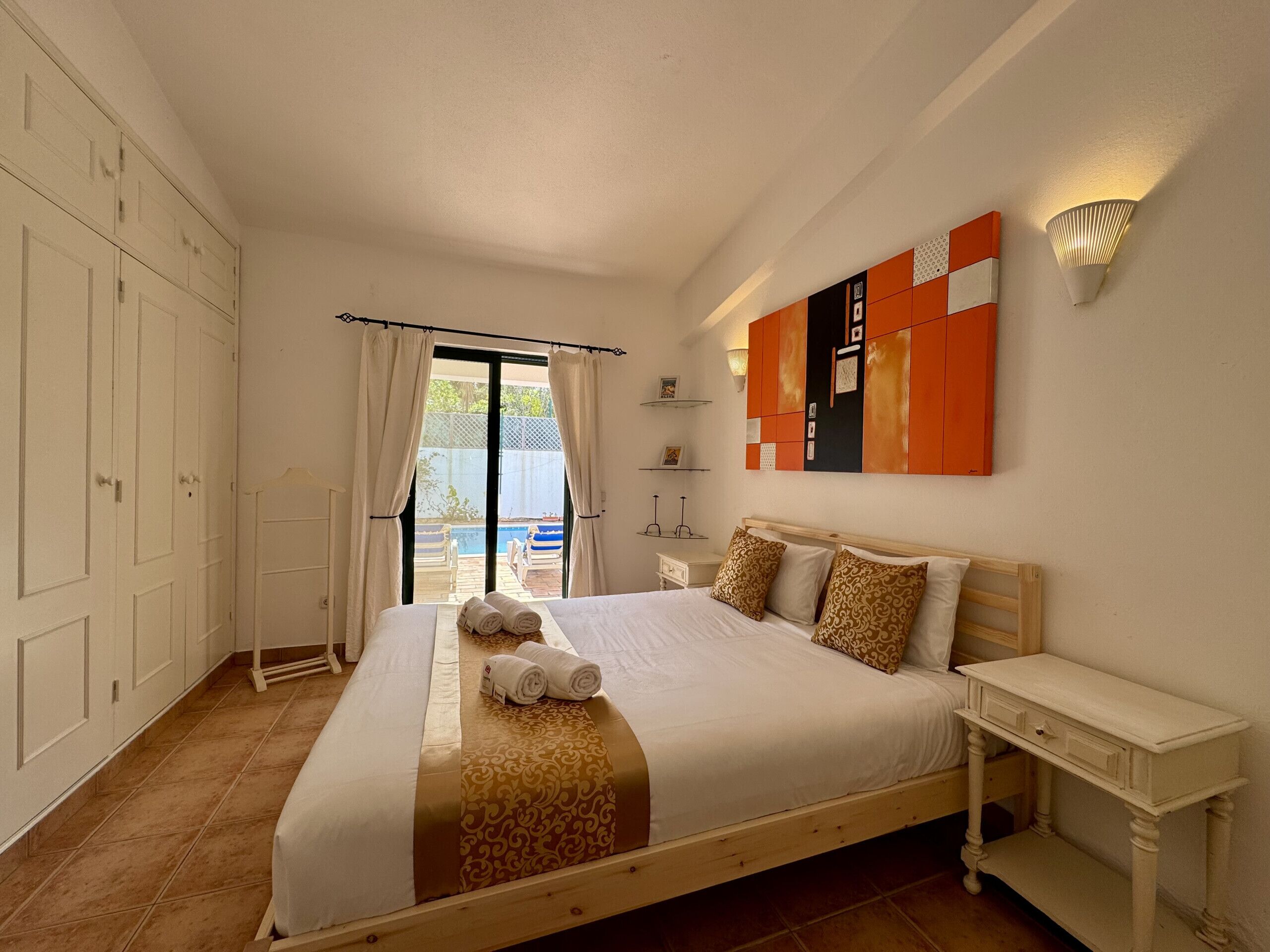 A bedroom with a bed, a nightstand, a wall-mounted light, a view of an outdoor pool, and a sliding door.