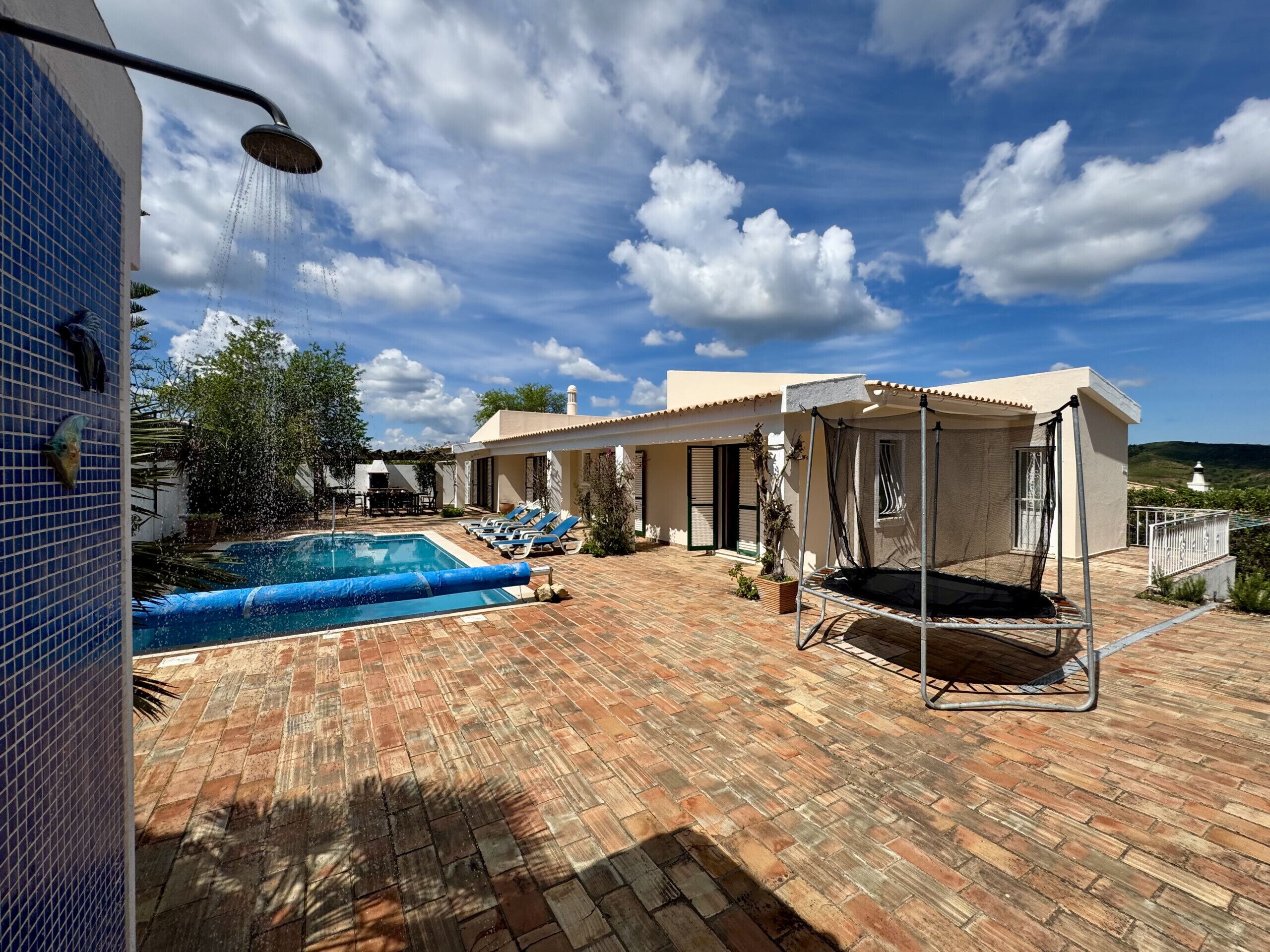 A pool area with a tiled wall, a covered seating area, and a view of a white building in the background.