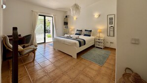 A bedroom with a bed, a chair, a bedside table, a lamp, a small shelf, and a view through a sliding door to an outdoor area. - Algarve Country Villa With Pool (Sao Bras de Alportel)