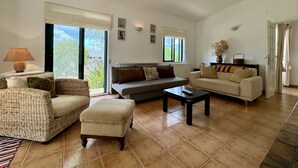 A living room with a sofa, ottoman, and coffee table, a lamp, and a view of greenery outside. - Algarve Country Villa With Pool (Sao Bras de Alportel)