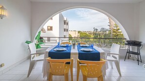 Espaço de refeições com mesa posta para dois, cadeiras brancas e uma churrasqueira.