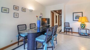 Uma área de refeições com mesa de vidro, cadeiras de vime e um vaso azul com flores.