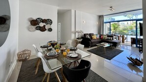Ein modernes Wohnzimmer mit einem Esstisch aus Glas, gedeckt für vier Personen, einer schwarzen Couch und einem großen Fenster mit Blick ins Freie.