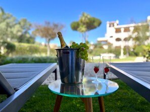 A metal bucket with a bottle of champagne, a bunch of grapes, and two glasses with strawberries on a glass table outdoors.
