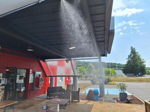 Uma área de estar coberta ao ar livre, com mesas e cadeiras, um edifício vermelho ao fundo e céu claro.