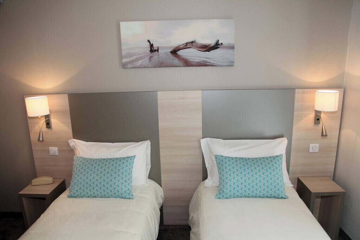 A hotel room with two beds, each with white bedding and teal patterned pillows. There are wall-mounted lamps on either side of the beds, and a painting of a beach scene above the beds.