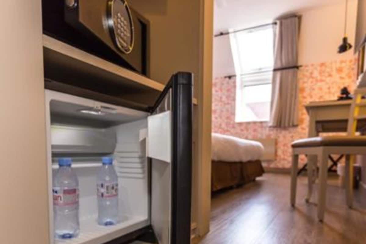 A compact hotel room with a small refrigerator containing bottled water, a bed with a white headboard, a wooden floor, and a window with curtains.