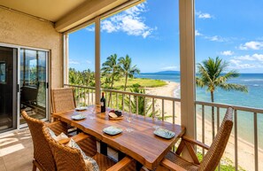 A wooden dining table with chairs, a bottle of wine, and a fruit platter, overlooking a beach with palm trees and the ocean. - Pure Luxury Kihei Rare 3BR Oceanfront Condo Freshly Redesigned Ocean Views (Kihei)