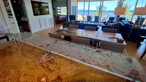 A modern kitchen with a glass-top island, a dining area with a long table, and a view of the ocean. - Pure Luxury Kihei Rare 3BR Oceanfront Condo Freshly Redesigned Ocean Views (Kihei)