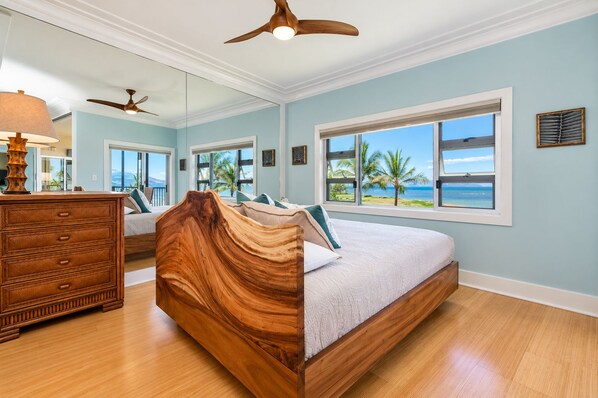 A bedroom with a wooden headboard, a bed with a white mattress, a dresser, and a large window with a view of the ocean. - Pure Luxury Kihei Rare 3BR Oceanfront Condo Freshly Redesigned Ocean Views (Kihei)