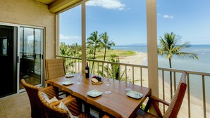 A balcony with a wooden table set for dining, overlooking a beach with palm trees and the ocean. - Pure Luxury Kihei Rare 3BR Oceanfront Condo Freshly Redesigned Ocean Views (Kihei)