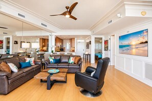 A spacious living room with a leather sofa, a coffee table, and a black armchair. There is a large painting on the wall and a ceiling fan. - Pure Luxury Kihei Rare 3BR Oceanfront Condo Freshly Redesigned Ocean Views (Kihei)
