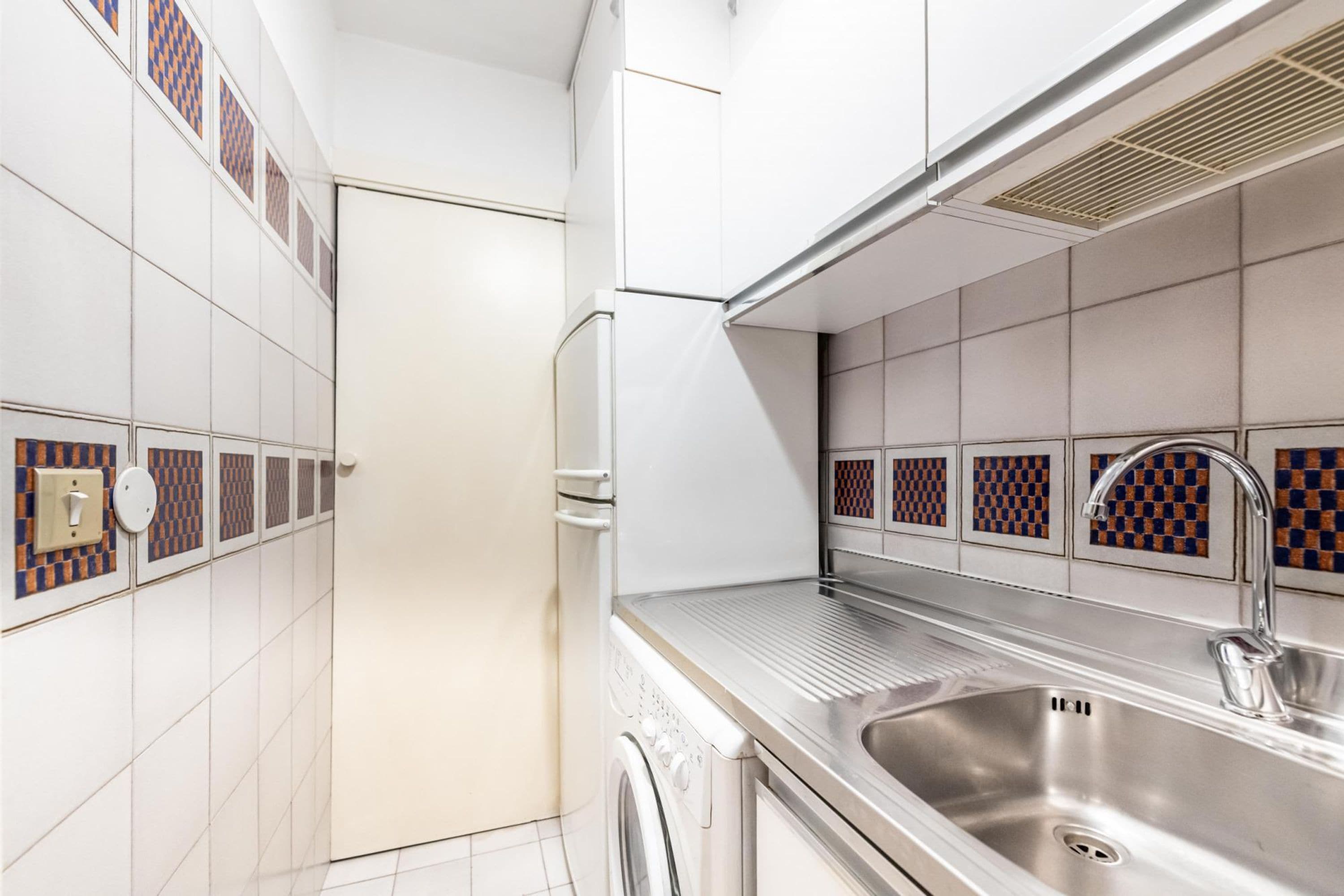 A compact kitchen with a stainless steel sink, a built-in washing machine, and patterned tile backsplash.