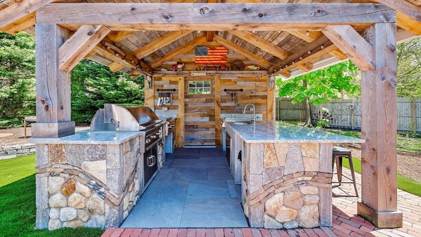 Une cuisine extérieure couverte, dotée de plans de travail en pierre et d’un barbecue, le tout entouré d’une pergola en bois.