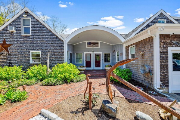 A house with a brick pathway leading to a front door, surrounded by greenery and a star decoration. - Luxury Cape Cod Compound-Heated Pool*Outdoor Kitchen*3 Connected Units (2302) (Eastham)