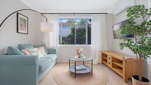 Una sala de estar moderna con un sofá azul claro, una mesa de centro redonda, un mueble de televisión de madera y un ventanal con cortinas.