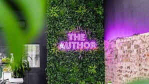 Acogedor interior de una cafetería con un letrero de neón que dice 'THE AUTHOR' en una pared verde, rodeado de plantas y una pared de ladrillo.