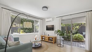 Una sala de estar moderna con un sofá, una mesa de centro, un televisor y un ventanal corredizo que da a un jardín.